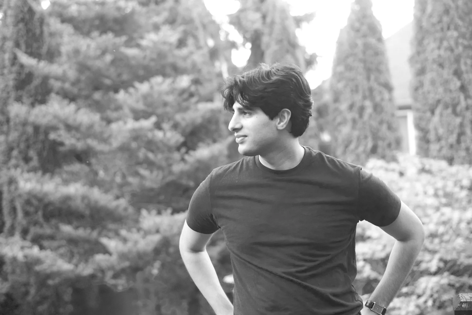 Rayyed Ahmed, standing outdoors among evergreens, looking off-frame — black and white portrait.